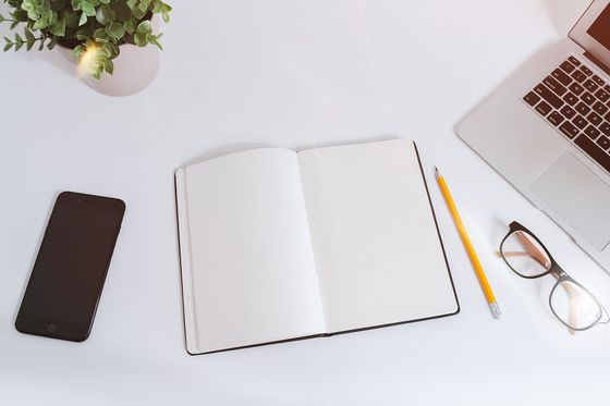Aufgeschlagenes leeres Notizbuch mit Bleistift zwischen Smartphone, Brille und Laptop auf weißem Tisch