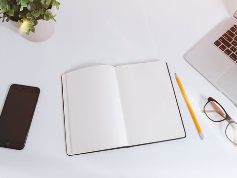Aufgeschlagenes leeres Notizbuch mit Bleistift zwischen Smartphone, Brille und Laptop auf weißem Tisch