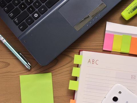 Laptop, Kugelschreiber, Haftnotizen und aufgeschlagenes Adressbuch auf einem Holzschreibtisch von oben