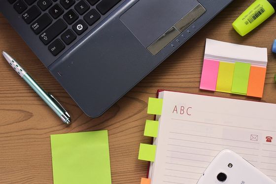 Laptop, Kugelschreiber, Haftnotizen und aufgeschlagenes Adressbuch auf einem Holzschreibtisch von oben